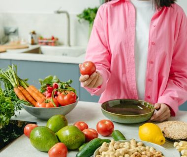Woman in pink overshirt holds tomato in kitchen with fresh fruits and veggies.